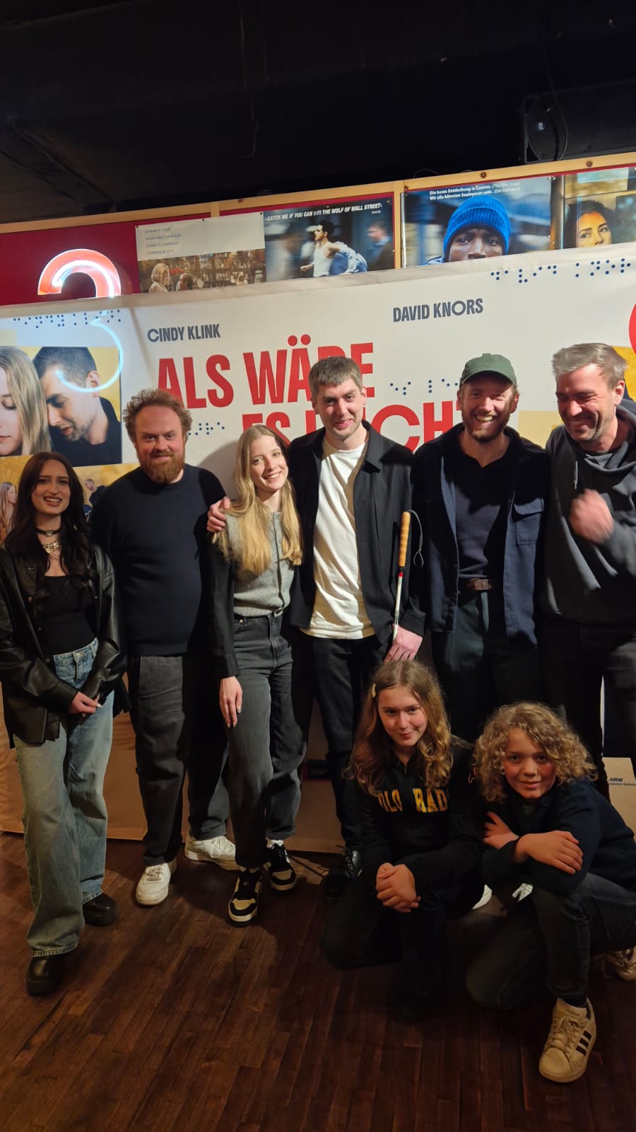 Gruppe von Menschen steht vor einer Wand mit dem Schriftzug 'ALS WÄRE ES LEICHT'.