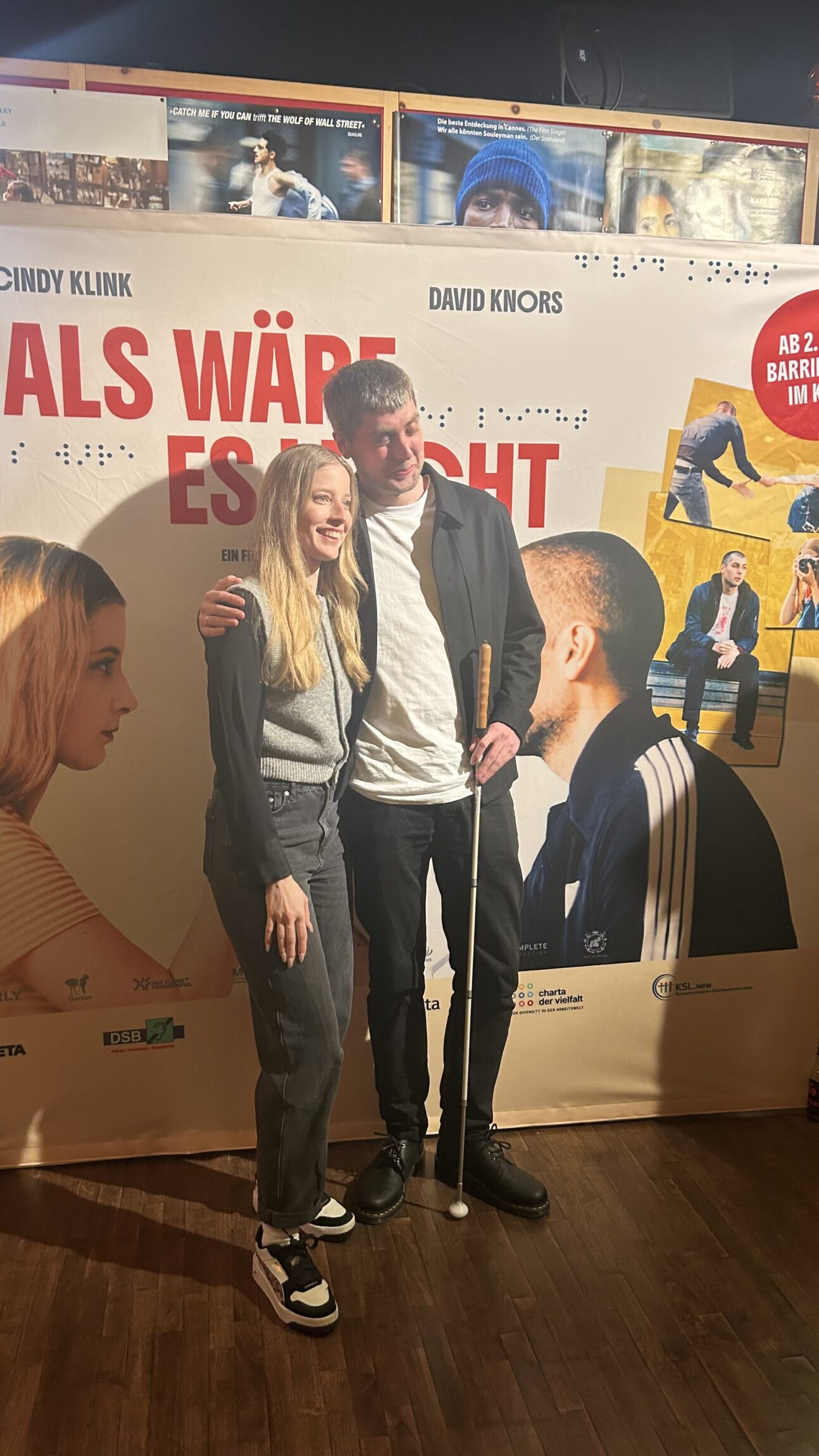 Zwei Personen stehen vor einer Wand mit einem Filmplakat zum Film Als wäre es leicht, die teilweise Text und Porträts zeigen.
