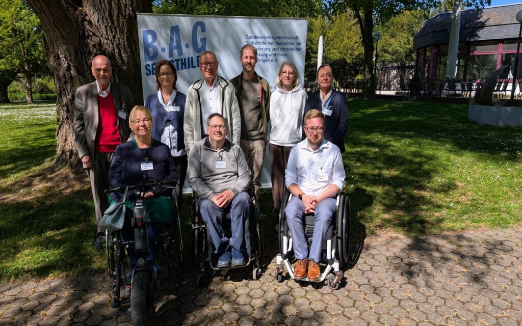 Gruppe von neun Personen, darunter zwei im Rollstuhl und eine mit Rollator, steht und sitzt vor einem Baum und einem Banner mit der Aufschrift 'B.A.G SELBSTHILFE' im Freien.