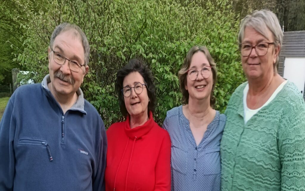 Vier Personen stehen nebeneinander vor grünen Büschen, zwei Männer und zwei Frauen, eine Frau trägt einen roten Pullover.