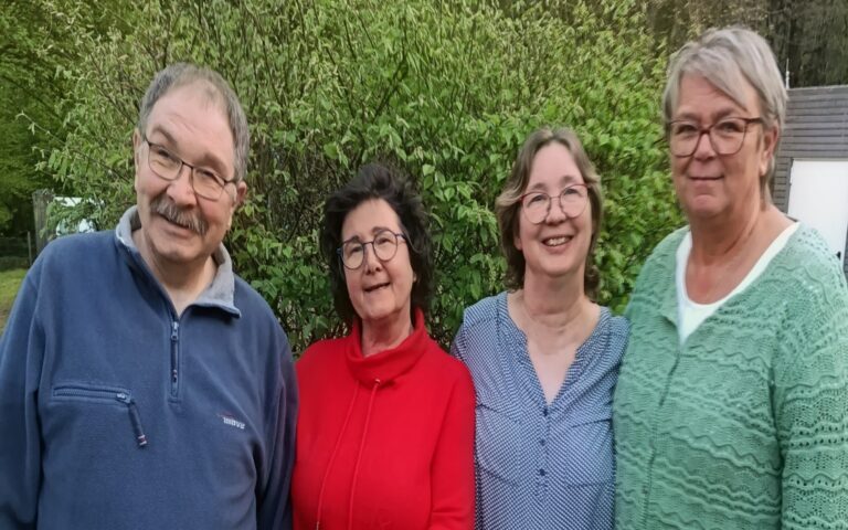 Vier Personen stehen nebeneinander vor grünen Büschen, zwei Männer und zwei Frauen, eine Frau trägt einen roten Pullover.