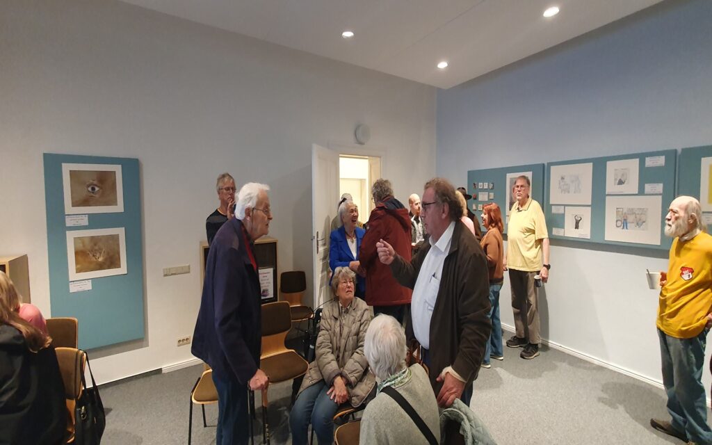 Mehrere ältere Menschen stehen und sitzen in einem Ausstellungsraum mit hellblauen Wänden. An den Wänden hängen gerahmte Zeichnungen oder Gemälde, die auf blauen Tafeln präsentiert werden. Einige Personen unterhalten sich, andere betrachten die ausgestellten Bilder. Die Atmosphäre wirkt ruhig und gesellig. Die Beleuchtung ist hell und gleichmäßig.