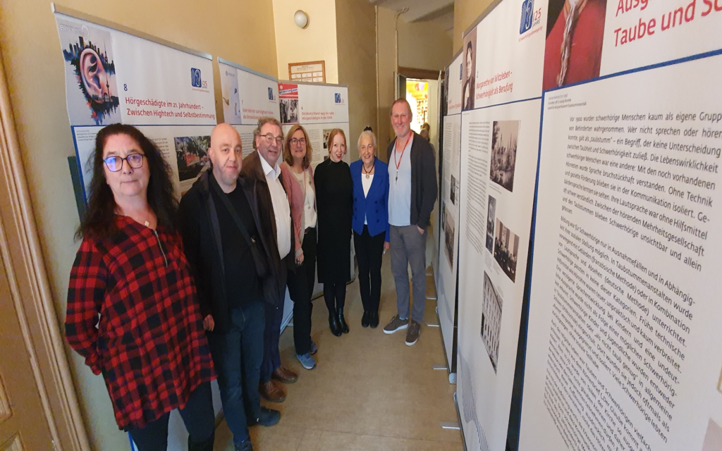 Mehrere Personen stehen in einem Flur zwischen zwei Reihen von Ausstellungstafeln. Die Tafeln zeigen großformatige Texte und Bilder, die thematisch aufeinander abgestimmt sind. Die Personen blicken zur Kamera, einige lächeln. Die Wände und der Boden sind hell, die Beleuchtung ist freundlich. Im Hintergrund ist eine Tür zu sehen.