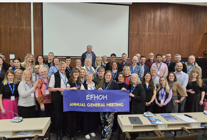 Eine große Gruppe von Menschen steht in einem Konferenzraum vor einer weißen Leinwand. Die meisten tragen Namensschilder an blauen Bändern um den Hals. Einige Personen in der vorderen Reihe halten ein lilafarbenes Banner mit der Aufschrift 'EFHOH ANNUAL GENERAL MEETING'. Im Vordergrund sind Tische mit Unterlagen und Getränken zu sehen. Die Wände sind holzvertäfelt.