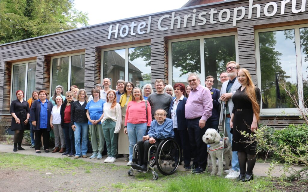 Gruppe von Menschen vor einem Gebäude mit der Aufschrift 'Hotel Christophorus', darunter eine Person im Rollstuhl und ein weißer Hund an der Leine.