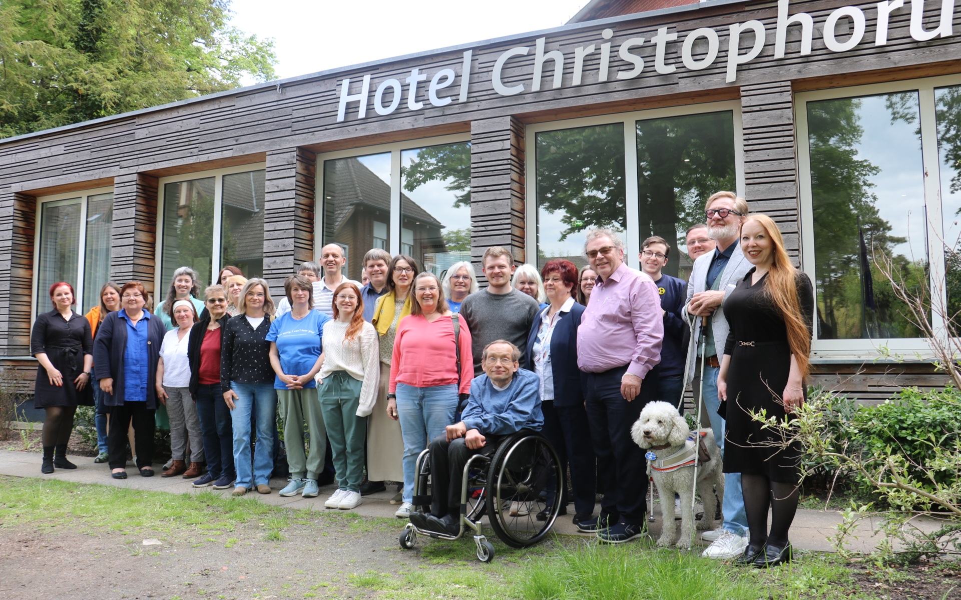 Gruppe von Menschen vor einem Gebäude mit der Aufschrift 'Hotel Christophorus', darunter eine Person im Rollstuhl und ein weißer Hund an der Leine.