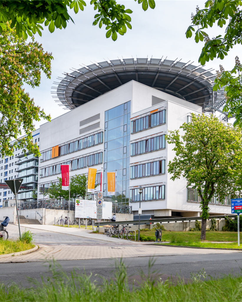 Ein mehrstöckiges, modernes Gebäude mit weißer Fassade und großen Glasflächen steht im Vordergrund. Auf dem Dach befindet sich eine auffällige, kreisförmige Konstruktion aus Metallstreben. Vor dem Gebäude sind mehrere bunte Fahnen an Masten befestigt. Bäume mit grünem Laub rahmen das Gebäude ein. Fahrräder stehen an einem Abstellplatz, und ein Gehweg führt am Gebäude vorbei. Im Vordergrund wächst Gras.