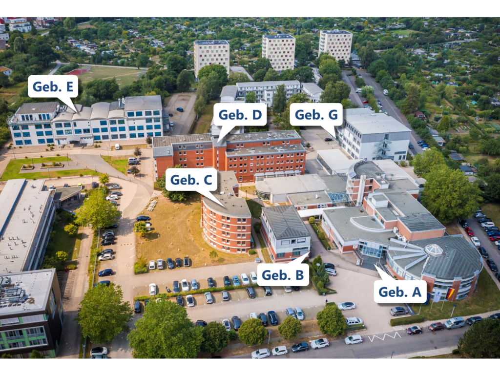 Luftaufnahme eines Gebäudekomplexes mit mehreren mehrstöckigen Gebäuden, die durch Wege und Parkplätze voneinander getrennt sind. Jedes Hauptgebäude ist mit einer weißen Sprechblase und schwarzer Beschriftung markiert: 'Geb. A', 'Geb. B', 'Geb. C', 'Geb. D', 'Geb. E' und 'Geb. G'. Im Hintergrund sind weitere Wohngebäude und viel Grün zu sehen. Autos stehen auf den Parkplätzen und an den Straßen rund um die Gebäude.