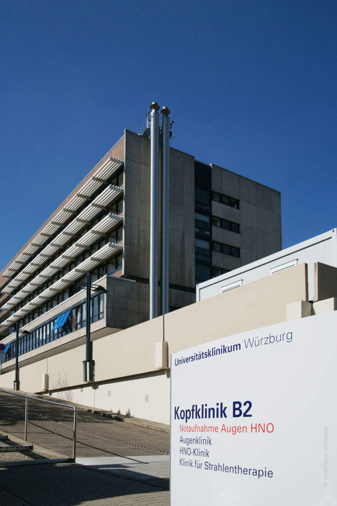 Ein mehrstöckiges, modernes Gebäude mit klaren Linien und großen Fensterfronten steht vor wolkenlosem, blauem Himmel. Im Vordergrund befindet sich ein weißes Schild mit blauer und roter Schrift. Darauf steht oben der Name eines Universitätsklinikums, darunter Hinweise zu verschiedenen Kliniken und Abteilungen, unter anderem zur Kopfklinik B2, Notaufnahme für Augen und HNO, HNO-Klinik und Klinik für Strahlentherapie. Das Gebäude wirkt funktional und ist von außen teilweise mit Metall- und Glaselementen verkleidet.