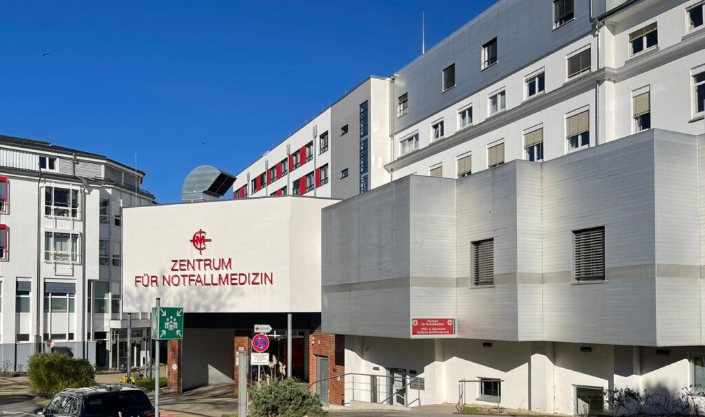 Ein mehrstöckiges, modernes Klinikgebäude mit weißer Fassade und zahlreichen Fenstern. Im Vordergrund befindet sich ein überdachter Eingangsbereich mit der Aufschrift 'Zentrum für Notfallmedizin' in roten Großbuchstaben. Links davon stehen Verkehrsschilder und ein grüner Wegweiser. Der Himmel ist wolkenlos und blau.