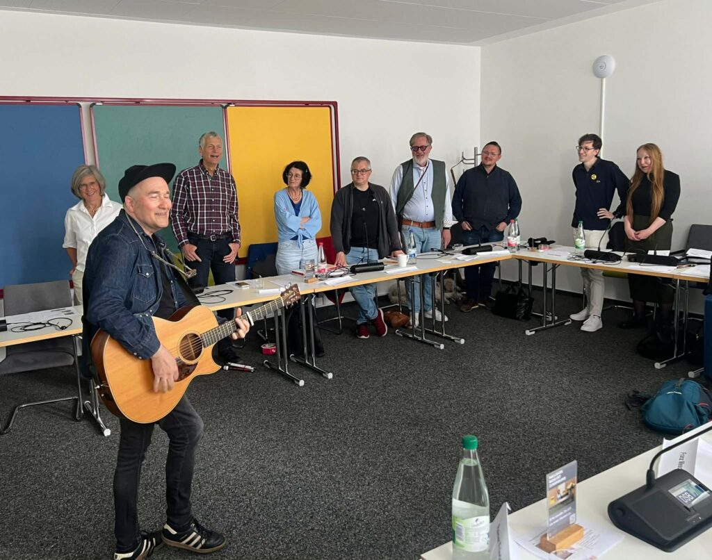 Person mit Gitarre steht vor einer Gruppe von acht Menschen, die in einem Raum mit Tischen und bunten Tafeln an der Wand stehen.