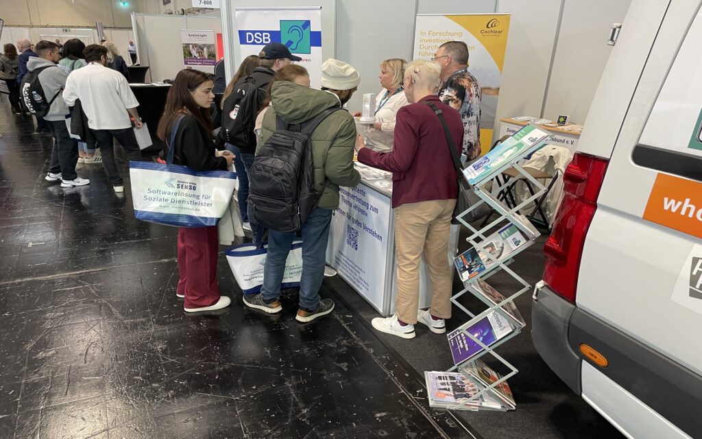 Mehrere Personen stehen an einem Messestand mit Informationsmaterial und einem Prospektständer neben einem weißen Fahrzeug.