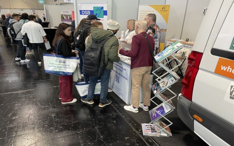 Mehrere Personen stehen an einem Messestand mit Informationsmaterial und einem Prospektständer neben einem weißen Fahrzeug.