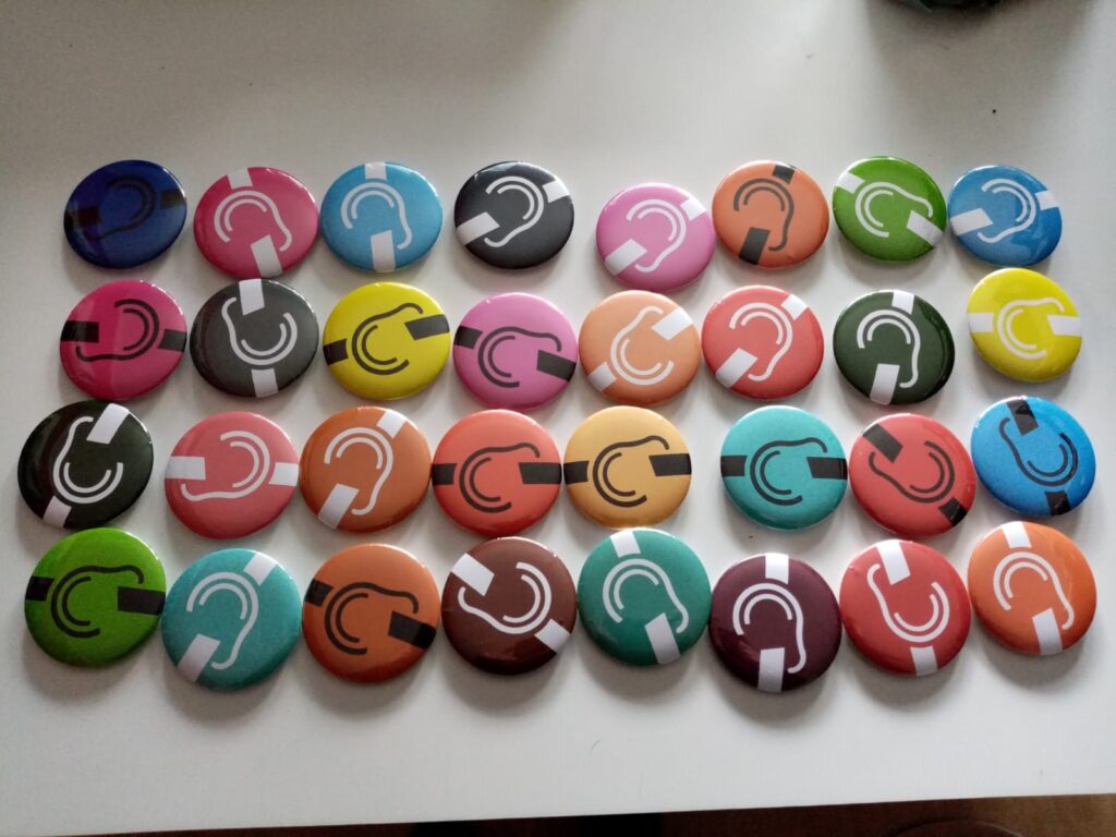 Mehrere bunte runde Buttons mit weißem durchgestrichenem weißen Ohr in verschiedenen Farben auf hellem Untergrund verteilt.