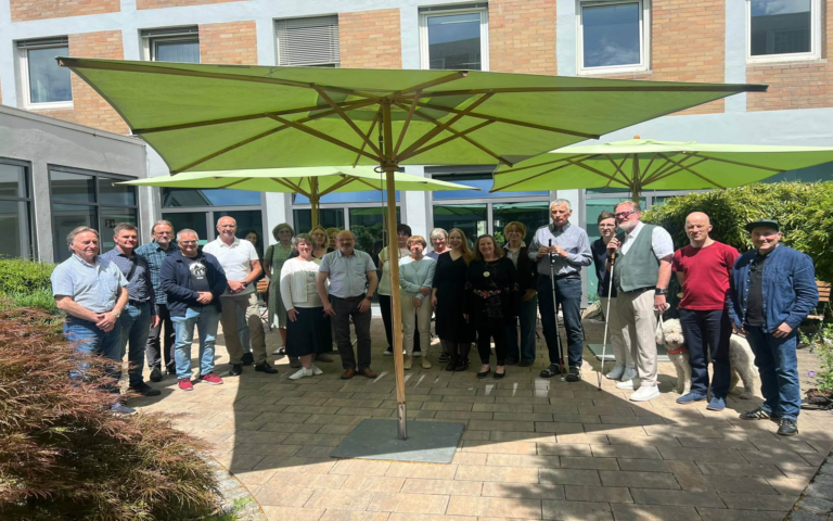 Eine Gruppe von Menschen steht im Halbkreis auf einer gepflasterten Terrasse vor einem modernen Gebäude. Im Hintergrund sind große, grüne Sonnenschirme aufgespannt. Die Personen tragen überwiegend Freizeitkleidung und blicken in Richtung Kamera. Die Szene ist von Tageslicht erhellt, einige Personen stehen im Schatten der Schirme. Im Vordergrund ist ein Teil eines Strauchs zu sehen.