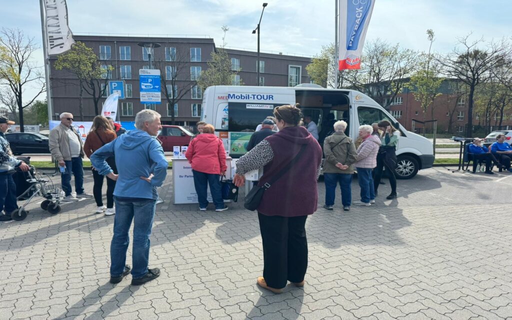 Mehrere ältere Menschen stehen auf einem gepflasterten Platz vor einem weißen Kleinbus mit der Aufschrift 'Mobil-TOUR'. Einige Personen unterhalten sich, andere schauen in Richtung des Fahrzeugs. Neben dem Bus steht ein kleiner Tisch mit Informationsmaterial. Im Hintergrund sind moderne Wohngebäude, einige Bäume und geparkte Autos zu sehen. Zwei hohe Fahnenmasten mit Werbefahnen ragen in den Himmel.