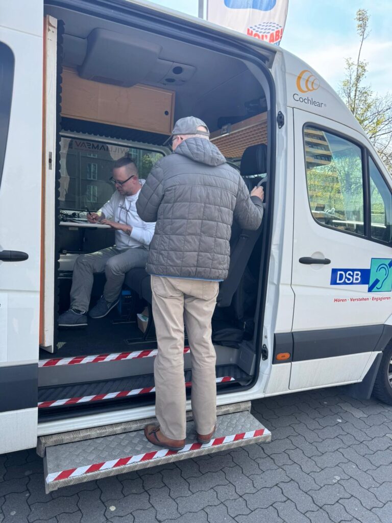 Ein Mann mit grauer Jacke und Mütze steht vor einem weißen Van mit offener Schiebetür, in dem eine Person sitzt.