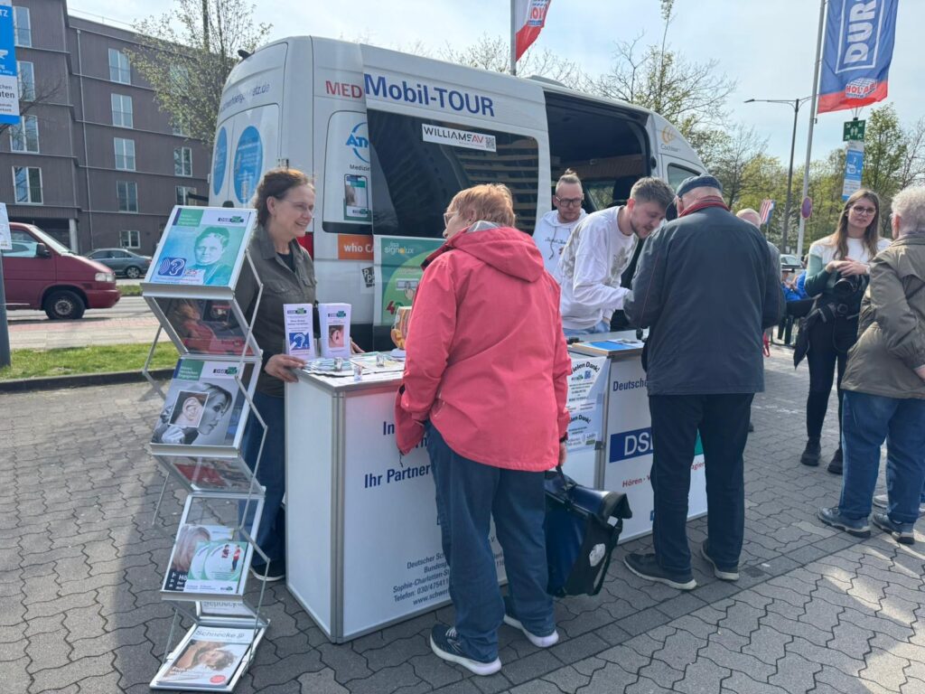 Menschen an einem Informationsstand vor einem weißen Transporter mit geöffneten Türen bei einem Aktionstag im Freien.