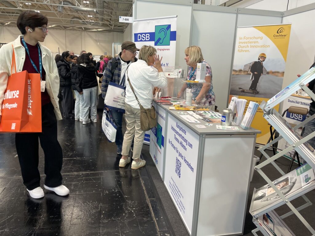 Mehrere Personen stehen an einem Messestand mit Informationsmaterial und Bannern im Hintergrund in einer Halle