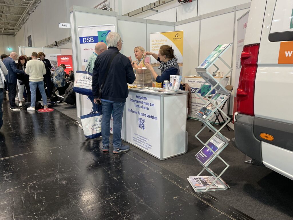 Messestand mit zwei Frauen und zwei Männern im Gespräch, neben einem Prospektständer und einem weißen Fahrzeug in einer Halle mit weiteren Ständen und Besuchern im Hintergrund.