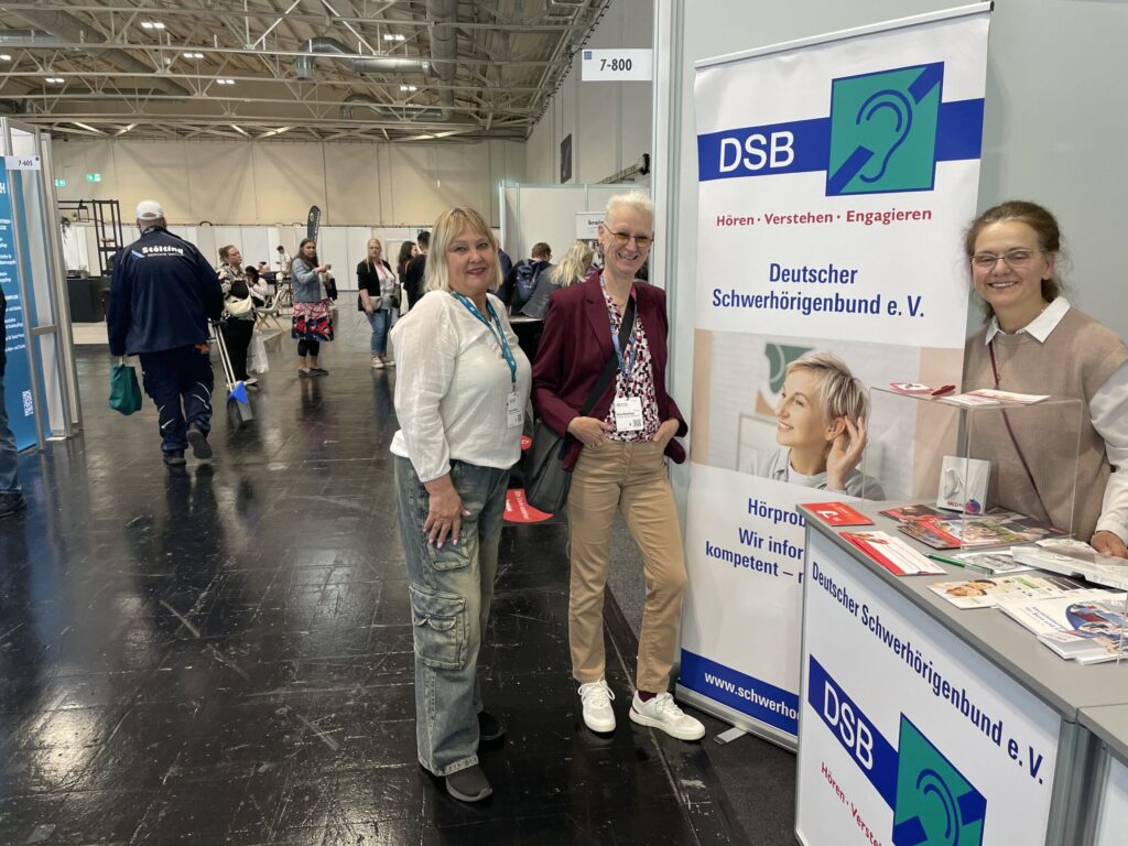 Messestand des Deutschen Schwerhörigenbunds mit Banner und Informationsmaterial auf einem Messestand in einer Halle mit mehreren Personen.