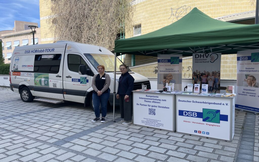 Zwei Frauen stehen vor einem weißen Van mit Aufschrift "DSB HörMobil-TOUR" und einem Informationsstand mit Bannern und Flyern des Deutschen Schwerhörigenbundes auf einem gepflasterten Platz.
