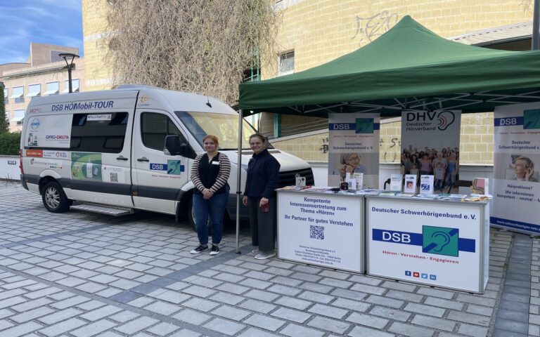 Zwei Frauen stehen vor einem weißen Van mit Aufschrift "DSB HörMobil-TOUR" und einem Informationsstand mit Bannern und Flyern des Deutschen Schwerhörigenbundes auf einem gepflasterten Platz.
