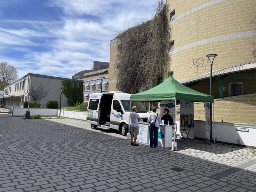 Ein weißer Transporter mit seitlicher Beschriftung steht neben einem grünen Pavillonzelt auf einem gepflasterten Platz. Zwei Personen stehen vor dem Zelt, eine weitere Person befindet sich dahinter. Der Hintergrund zeigt ein rundes, gelbliches Gebäude mit rankenden Pflanzen an der Fassade sowie weitere moderne Gebäude. Der Himmel ist leicht bewölkt.