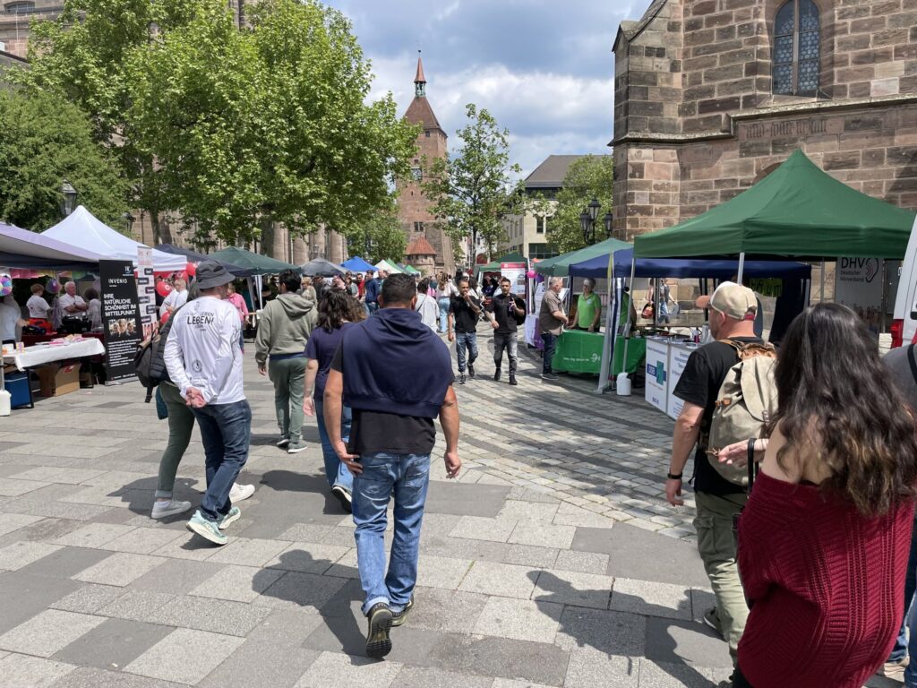 Menschen gehen an Ständen auf einem gepflasterten Platz entlang, im Hintergrund Bäume und historische Gebäude.