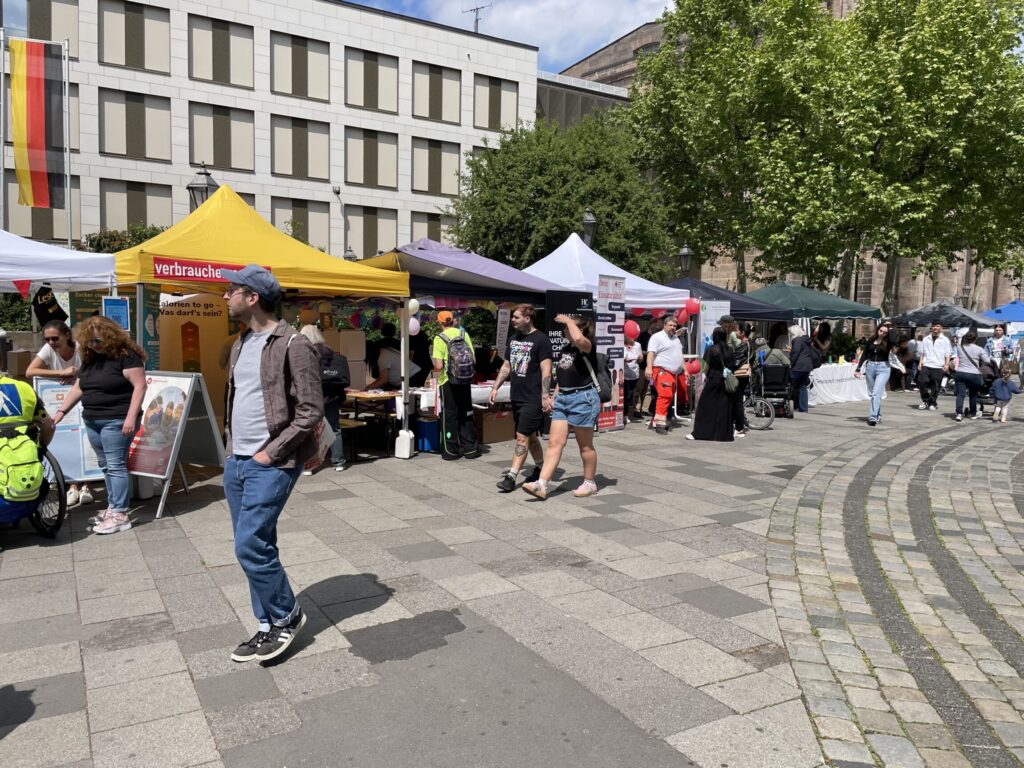 Marktstände mit bunten Zelten und Menschen auf gepflastertem Platz vor einem modernen Gebäude mit deutscher Flagge.