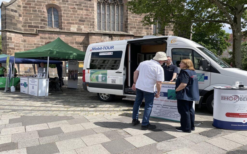 Zwei Personen stehen vor einem weißen Informationsstand der DSB HÖRMobil-TOUR auf einem gepflasterten Platz vor einer Kirche, daneben grüne und blaue Pavillons mit Informationsständen