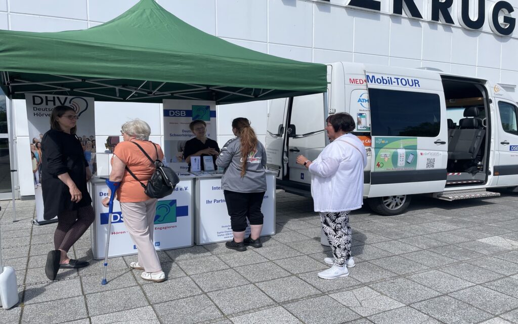 Menschen stehen vor einem grünen Pavillon mit Informationsstand und einem weißen Van mit verschiedenen Werbeaufklebern von Firmen.