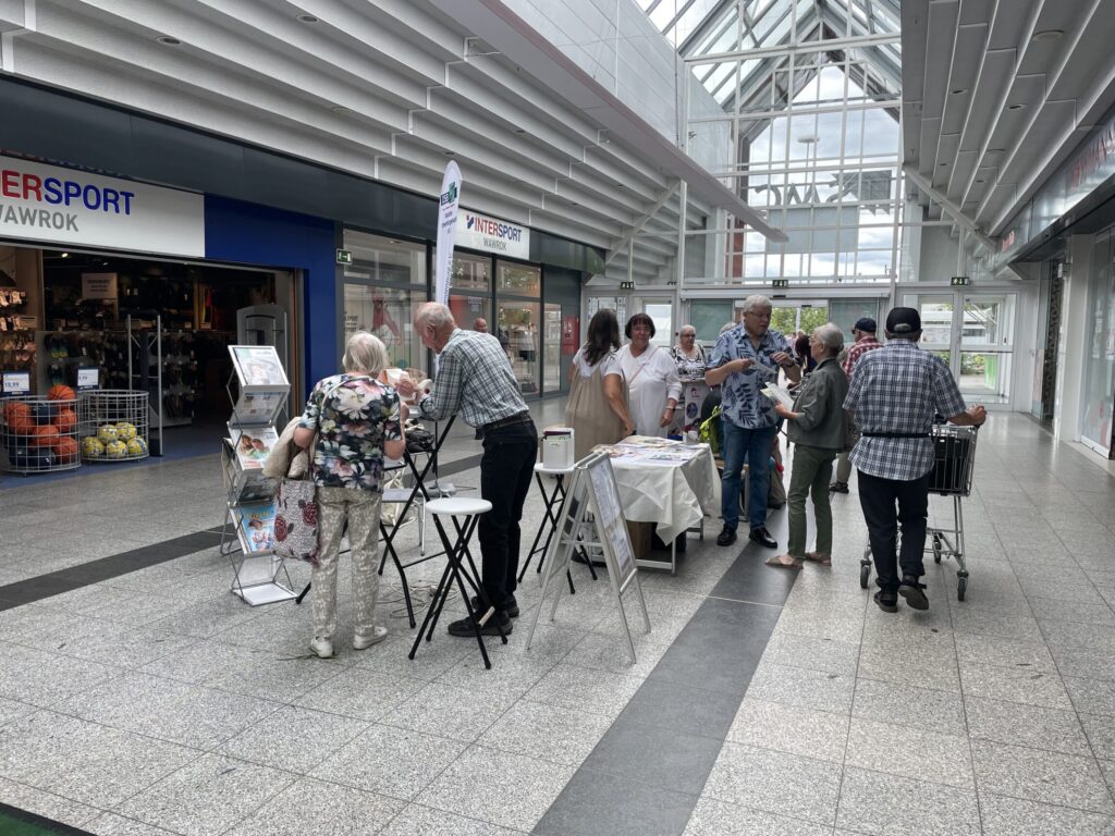 Gruppe von Menschen steht und spricht in einem hellen Einkaufszentrum mit Glasdach und Verkaufsständen.