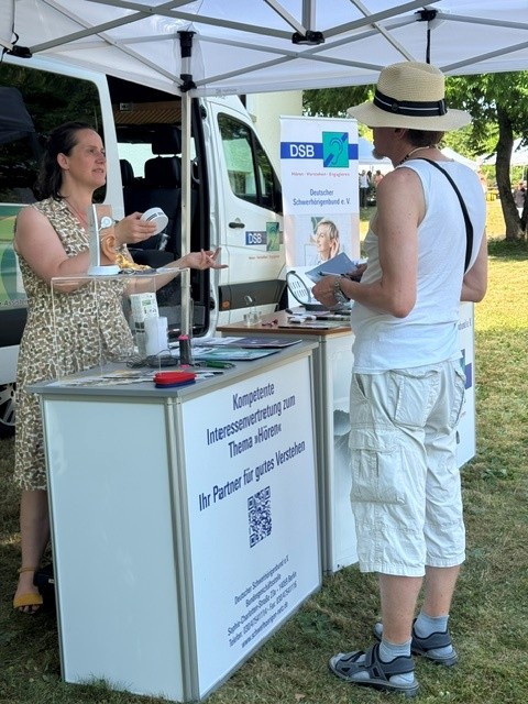 Im Vordergrund steht ein Informationsstand mit weißer Theke, auf der verschiedene Gegenstände liegen. Eine Frau mit langen dunklen Haaren, hellem Oberteil und Rock steht hinter der Theke und spricht mit einer Person, die einen hellen Sonnenhut, ein ärmelloses weißes Oberteil und helle Shorts trägt. Im Hintergrund ist ein weißer Transporter mit geöffneten Türen zu sehen. Über dem Stand ist ein weißes Pavillondach gespannt. Auf der Theke ist ein Text zum Thema Hören und ein QR-Code zu erkennen. Ein Plakat mit dem Logo des Deutschen Schwerhörigenbundes und weiteren Informationen ist an der Seite angebracht. Die Szene spielt im Freien auf einer Wiese bei sonnigem Wetter.