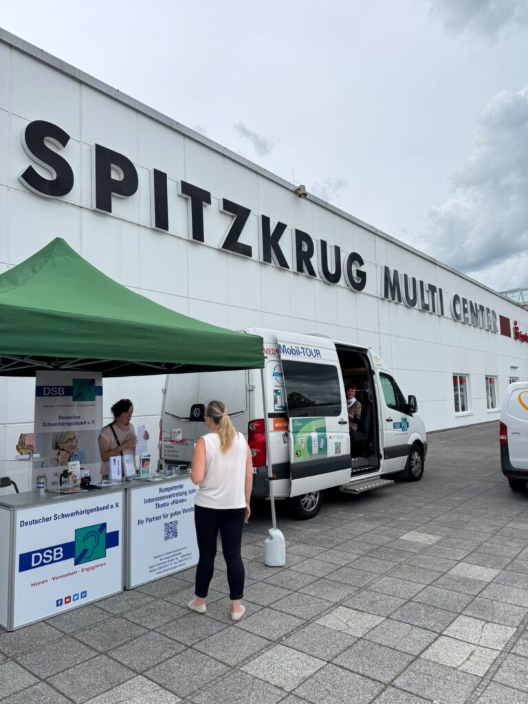 Außenansicht des SPITZKRUG MULTI CENTER mit einem Informationsstand des Deutschen Schwerhörigenbundes. Zwei Personen stehen am Stand.