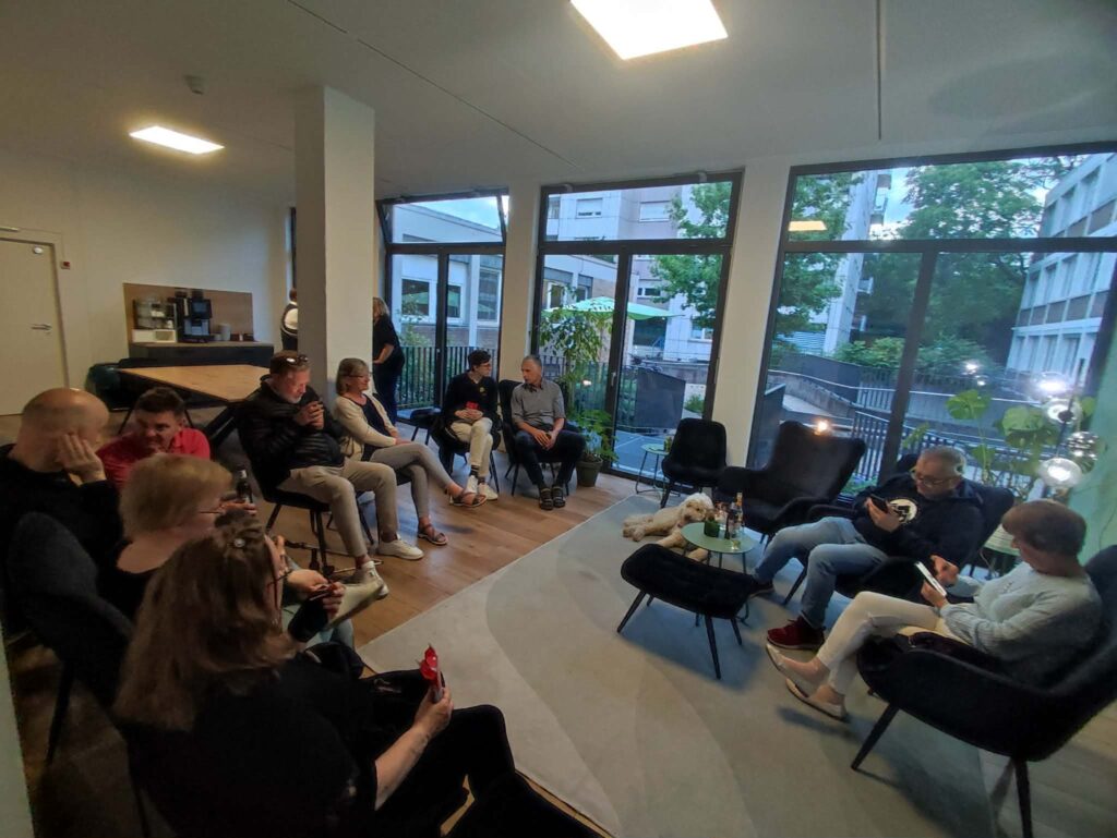 Mehrere Personen sitzen in einem modernen, hellen Raum mit großen Fenstern, die den Blick auf ein städtisches Gebäude und Bäume freigeben. Die Sitzgruppe besteht aus dunklen Sesseln, die im Kreis um einen niedrigen Glastisch angeordnet sind. Im Hintergrund steht eine Kaffeemaschine auf einer Theke. Einige Personen halten Getränke in der Hand, andere schauen auf ihre Smartphones. Die Atmosphäre wirkt entspannt und informell.
