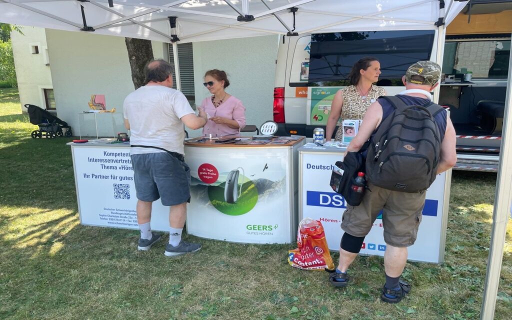 Zwei Frauen stehen hinter einem Informationsstand im Freien, der unter einem weißen Pavillon aufgebaut ist. Vor dem Stand unterhalten sich zwei Männer mit den Frauen. Auf den Tischen liegen verschiedene Broschüren und Gegenstände. Im Hintergrund ist ein Transporter mit geöffneter Tür zu sehen. Die Szene spielt auf einer Wiese bei sonnigem Wetter.