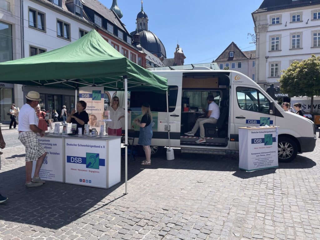 Marktstand mit grünem Pavillon und Informationsständen des Deutschen Schwerhörigenbunds vor einem weißen Fahrzeug auf gepflastertem Platz in einer Stadt mit historischen Gebäuden.