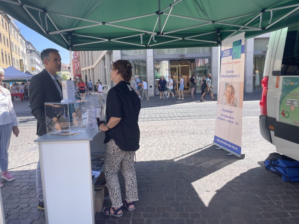 Zwei Personen unter einem grünen Pavillon an einem Informationsstand auf einem gepflasterten Platz mit Passanten im Hintergrund.