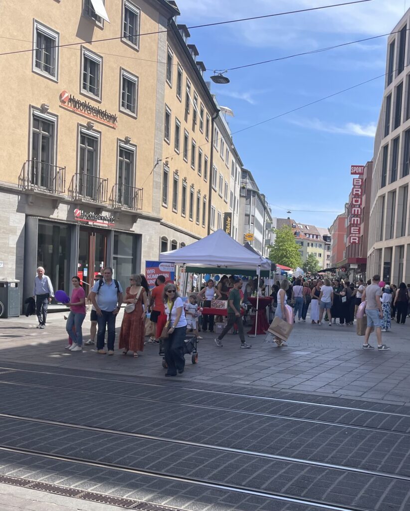 Belebte Fußgängerzone mit Informationsstand unter Pavillon und Menschen vor historischen Gebäuden.