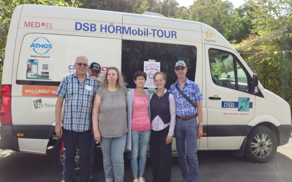 Ein weißer Kleinbus mit der Aufschrift 'DSB HÖRMobil-TOUR' steht im Freien. An der Seite des Fahrzeugs sind mehrere Logos und Schriftzüge angebracht, darunter 'MED-EL', 'ATMOS Medizintechnik', 'Cochlear' und 'DSB Deutscher Schwerhörigenbund'. Vor dem Bus stehen fünf Erwachsene nebeneinander, einige mit Sonnenbrille oder Brille. Sie tragen legere Kleidung und blicken in Richtung Kamera. Im Hintergrund sind Bäume und ein Zaun zu sehen.
