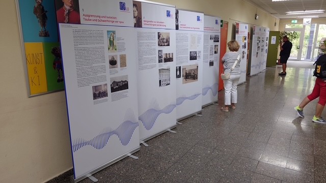Mehrere große, freistehende Informationstafeln mit Texten und historischen Fotografien stehen in einem Flur mit glänzendem Boden. Die Tafeln zeigen unten eine wellenförmige Grafik in Blautönen. Eine Person mit heller Kleidung betrachtet eine der Tafeln aus nächster Nähe. Im Hintergrund sind weitere Personen und zusätzliche Tafeln zu sehen. An der Wand links hängt ein farbiges Plakat mit dem Schriftzug 'KUNST & KI'.