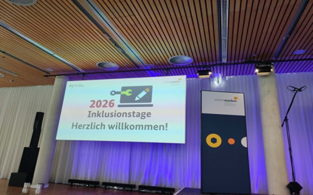 Leinwand mit der Projektion '2026 Inklusionstage Herzlich willkommen' in einem Saal mit Holzdecke und Bühnenbeleuchtung.