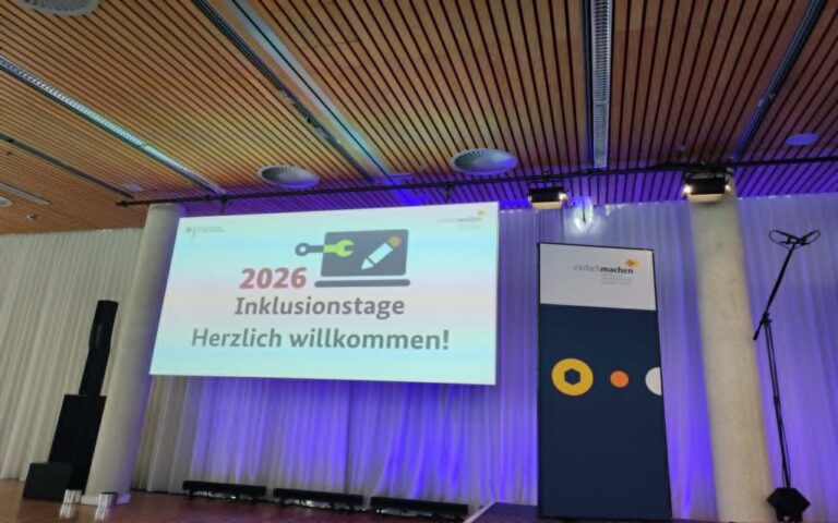 Leinwand mit der Projektion '2026 Inklusionstage Herzlich willkommen' in einem Saal mit Holzdecke und Bühnenbeleuchtung.