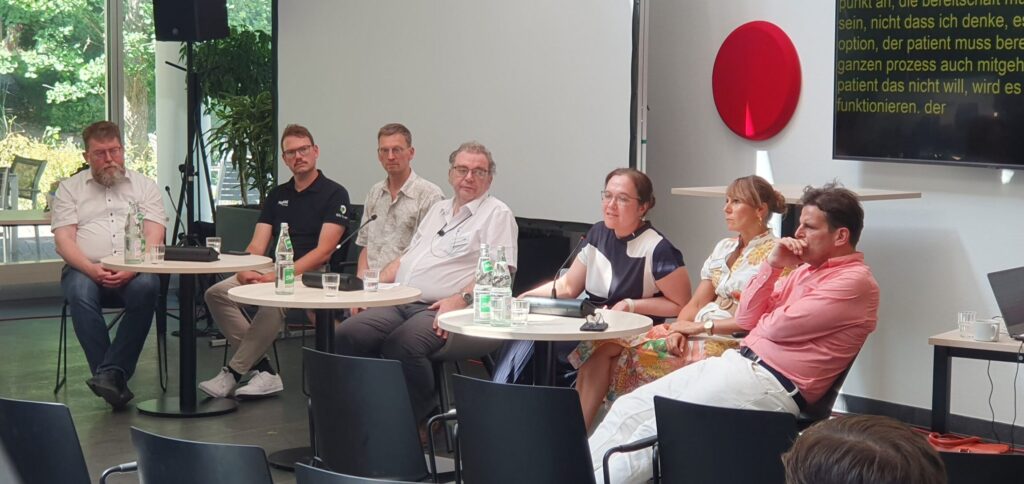 Sechs Personen sitzen nebeneinander auf einem Podium in einem hellen Raum. Vor ihnen stehen kleine runde Tische mit Wasserflaschen und Gläsern. Im Hintergrund ist eine große Fensterfront mit Blick ins Grüne zu sehen. Rechts an der Wand hängt ein roter Kreis als dekoratives Element. Daneben befindet sich ein Bildschirm mit gelber Schrift auf schwarzem Grund, der Untertitel anzeigt. Die Personen tragen unterschiedliche, überwiegend helle Kleidung und blicken in verschiedene Richtungen. Einige haben Notizbücher oder Unterlagen vor sich liegen.