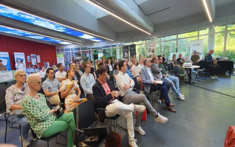 Ein Konferenzraum mit zahlreichen sitzenden Personen, die nach vorn blicken. Die Teilnehmenden tragen überwiegend legere bis businessmäßige Kleidung. Im Hintergrund sind große Fensterfronten mit Blick ins Grüne sowie mehrere Plakate an einer roten Wand zu sehen. Einige Personen halten Notizblöcke oder Laptops, während andere aufmerksam zuhören. Die Atmosphäre wirkt ruhig und konzentriert.