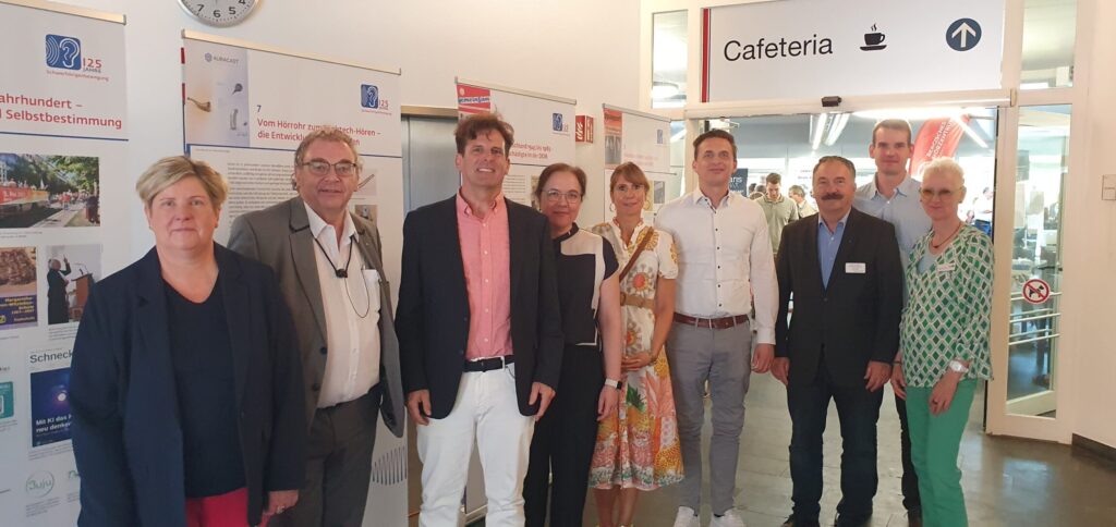 Eine Gruppe von Menschen steht nebeneinander in einem Flur vor einer Wand mit Plakaten und Informationstafeln. Im Hintergrund ist ein Schild mit der Aufschrift 'Cafeteria' und Symbolen für eine Tasse und Besteck zu sehen. Die Personen tragen überwiegend formelle Kleidung, darunter Anzüge, Blazer und ein gemustertes Kleid. Die Stimmung wirkt freundlich und die Personen blicken in Richtung Kamera.