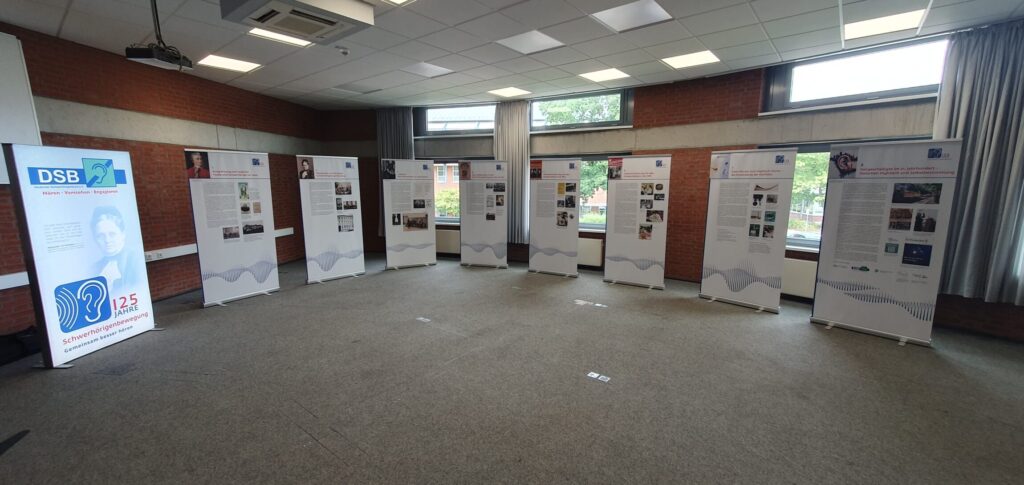 Mehrere Ausstellungstafeln stehen in einem großzügigen, hellen Raum mit Teppichboden, abgehängter Decke und großen Fenstern. Die Tafeln sind in einem leichten Bogen aufgestellt und zeigen verschiedene Texte, Fotos und Grafiken. Links steht ein beleuchteter Aufsteller mit einem Porträtfoto, einem Logo und weiteren grafischen Elementen. Tageslicht fällt durch die Fenster, die Wände sind teilweise mit roten Klinkern verkleidet.