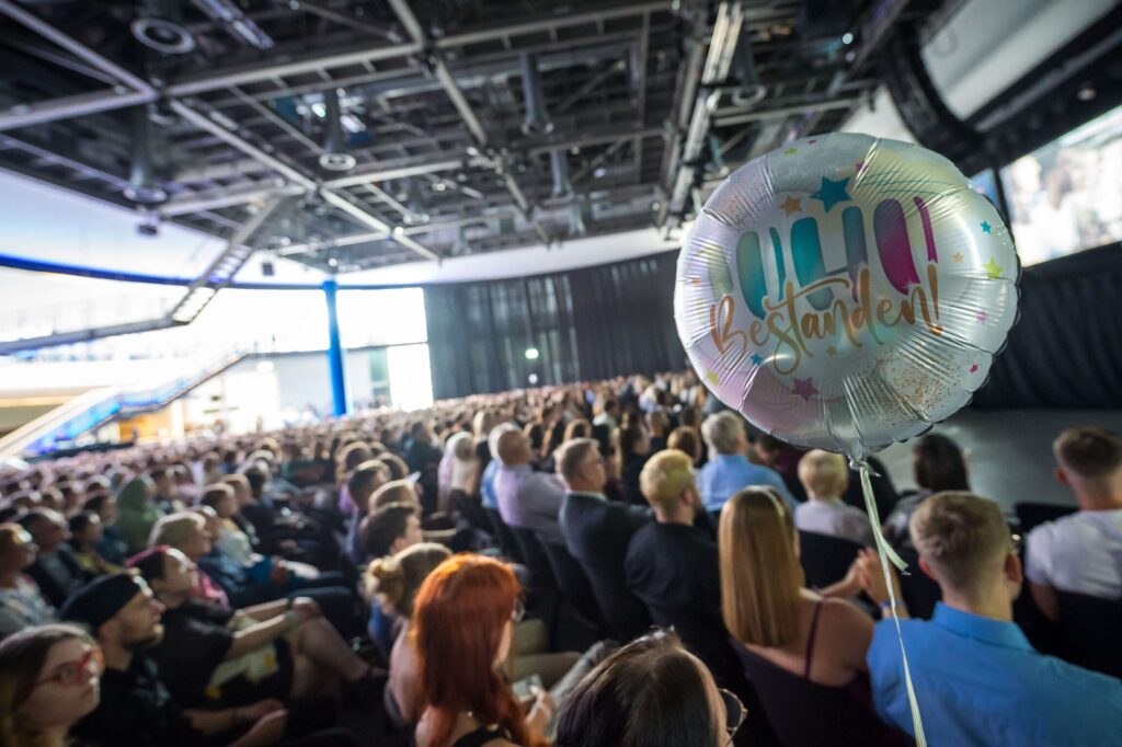 m Vordergrund schwebt ein silberner Folienballon mit buntem Aufdruck und Sternmotiven, auf dem das Wort 'bestanden!' steht. Im Hintergrund sitzt eine große Menschenmenge in einem modernen Saal mit hoher Decke und sichtbaren Metallträgern. Die Personen blicken in Richtung einer Bühne die am rechten Bildrand angeschnitten ist. Tageslicht fällt durch große Fenster an der linken Seite des Raumes.