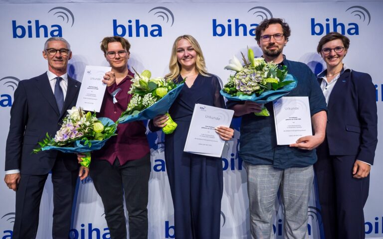 Fünf Personen stehen vor einer Wand mit dem Schriftzug 'biha', drei von ihnen halten Urkunden und Blumensträuße.